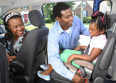 Father securing his young daughter in a booster seat, while the mother looks on from the passenger seat.