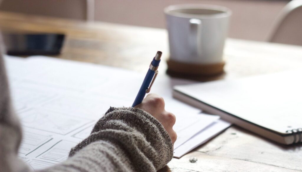 person's hand holding a pen with study materials and coffee mug on a desk