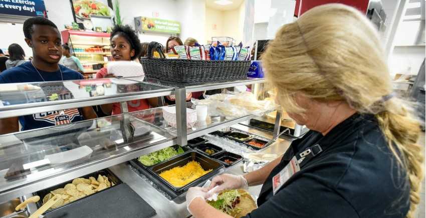 A food service worker in gloves prepares a taco for students in a school cafeteria line, with fresh ingredients like lettuce, cheese, and chips visible on the counter.