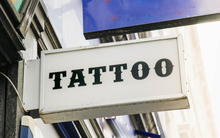 A white rectangular sign with the word 'TATTOO' in bold black letters, mounted on a building facade.