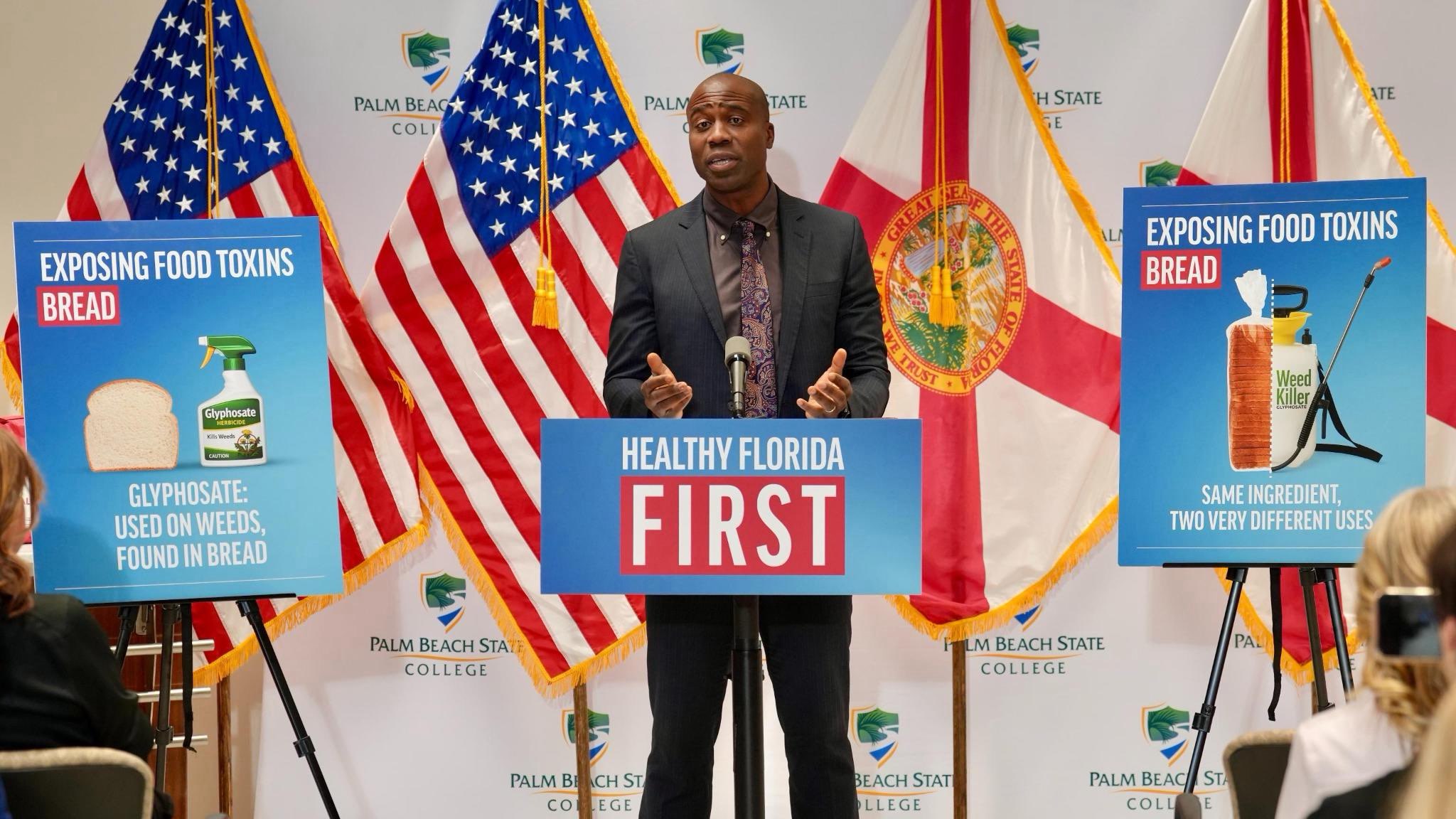 A man speaks at a podium with 'Healthy Florida First' on it, flanked by signs warning about glyphosate (weed killer) found in bread, with American and Florida flags in the background.
