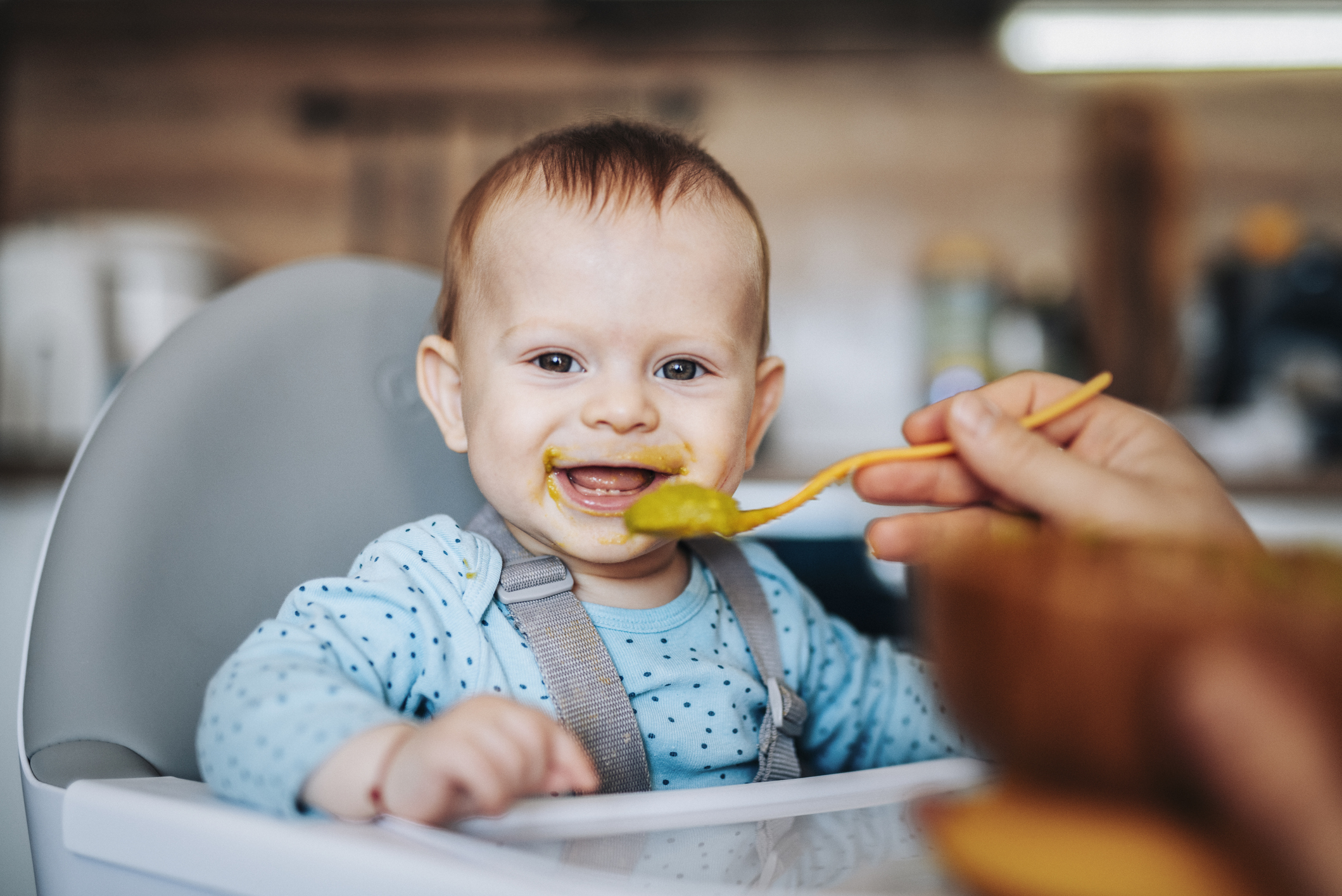 A smiling baby with food smeared around their mouth sits in a high chair while being fed from a spoon.
