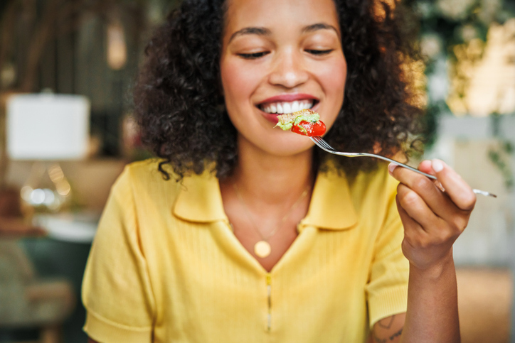 A smiling woman eating a cherry tomato with green topping from a fork