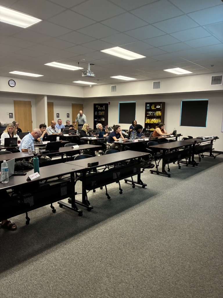 A classroom filled with adults seated at long tables, attending a workshop. A projector is on the ceiling, and shelves with yellow and black instruments are on the front wall.