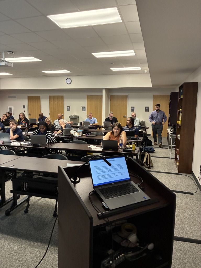 A classroom scene with multiple people seated at desks, looking towards a presentation. A laptop on a podium displays a slide about 'Exercising Violations' and regulatory compliance. A man stands in the back, observing the group.