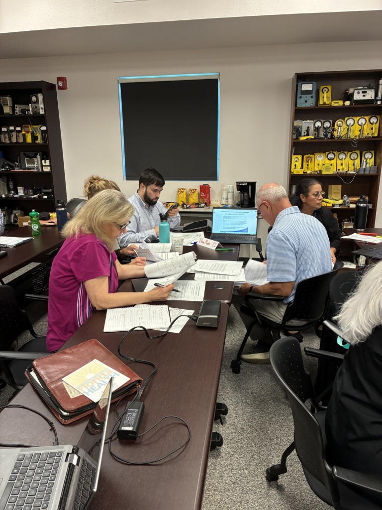 A group of people seated around a conference table, looking at laptops and papers. A bookshelf filled with yellow radiation detection devices is in the background.