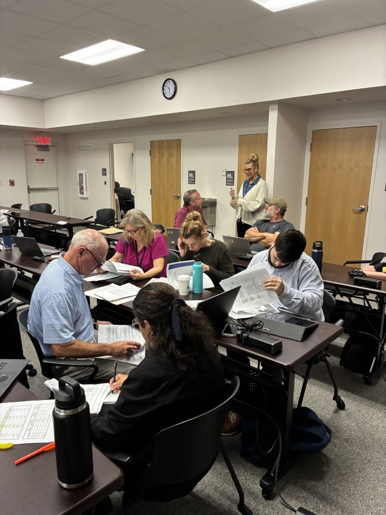 A group of adults seated at individual desks in a classroom, looking at papers and laptops during a training session. One woman stands and gestures while talking to a seated man.