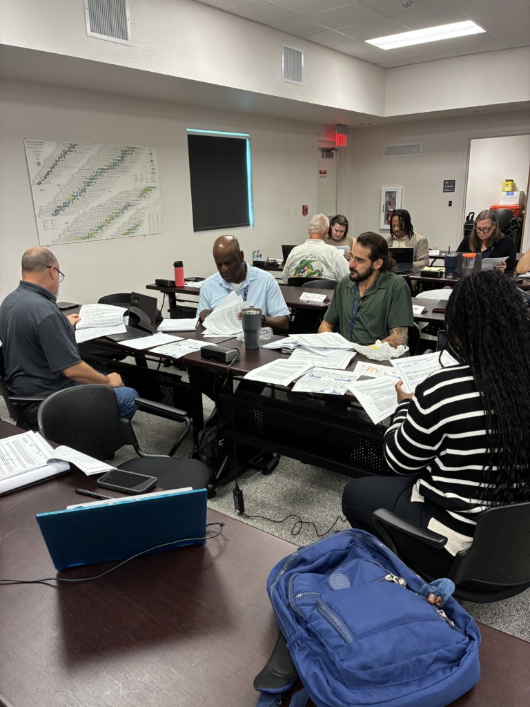 ```json { "description": "The image captures a diverse group of individuals actively engaged in what appears to be a training or educational session within a classroom setting. The perspective is from a slightly elevated, side angle, looking down a row of desks. Participants are seated at individual dark brown desks, each covered with various papers and documents, suggesting focused study or work. nnIn the foreground, a person with short hair and glasses, wearing a dark grey t-shirt, is intently focused on the papers before them. Moving further into the scene, a Black man in a light blue polo shirt is seen reviewing documents, with a dark grey travel mug on his desk. Next to him, a man with a beard and long dark hair, wearing a green shirt and a blue lanyard, is also engrossed in papers, with a white paper bag (possibly for food) nearby and a desk nametag reading 'Paul-FL'. Behind him, a person with dark braids or dreadlocks, wearing a black and white striped long-sleeved sweater, is similarly focused on documents. A blue backpack rests on the carpeted floor beside their chair. Further down the rows, other participants are visible, including one wearing a white shirt with 'Gainesville Fire Rescue' visible on the back, all appearing to be studying or working.nnThe room features light-colored walls, bright overhead fluorescent lighting, and a grey carpeted floor. On the left wall, a large 'Chart of the Nuclides' is prominently displayed, suggesting a scientific or technical subject matter. A dark screen or whiteboard with a blue glow is also visible on the far wall. An 'EXIT' sign with a red light, a 'STORAGE 139' sign on a door, and a 'DO NOT BLOCK' sign are visible on the walls, along with a framed picture. The overall atmosphere conveys a focused and productive learning environment.", "caption": "Focused learning in action! Participants, including members of Gainesville Fire Rescue, are deeply engaged in a training session, reviewing documents at their desks. A 'Chart of the Nuclides' hints at the technical nature of the subject.", "altText": "A diverse group of people seated at desks in a classroom, intently reviewing papers during a training session. A large 'Chart of the Nuclides' is visible on