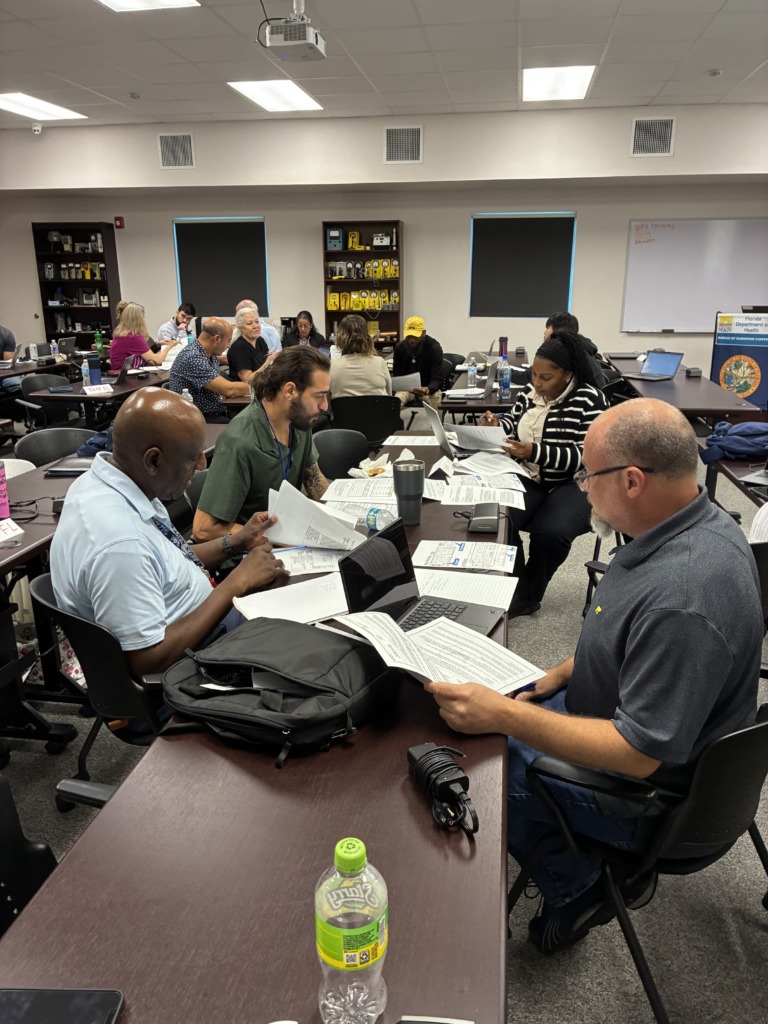 A diverse group of people attending a training session in a classroom, focused on papers and laptops at long tables, with 'Florida Department of Health, Bureau of Radiation Control' signage visible on the wall.