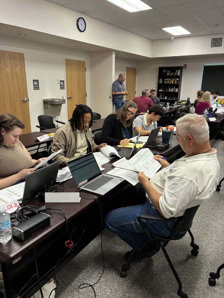 ```json { "description": "The image depicts an active indoor work or training session, likely within an emergency operations center or a similar command-and-control environment. The perspective is slightly elevated, looking down at multiple individuals seated at long, dark brown tables arranged in rows across a gray carpeted floor. nnIn the immediate foreground on the right, a man with short gray hair, wearing a white polo shirt with 'EM' (likely for Emergency Management) on the sleeve and blue jeans, is intently focused on papers he holds in his hands, with a pen poised as if filling out a form. A silver laptop is open on the table in front of him. nnMoving left and further into the frame, several other participants are visible. A woman with glasses and long dark hair is seated, looking at a laptop and papers, with a yellow notepad nearby. Next to her, a person with dreadlocks, wearing a light-colored jacket, is also engrossed in work on a laptop and documents. Another