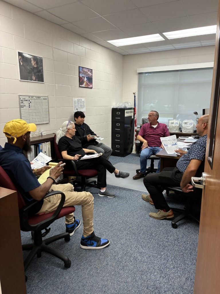 A group of five people in an office, seated and reviewing documents, with an 'IN/OUT PLANNER' board on the wall.