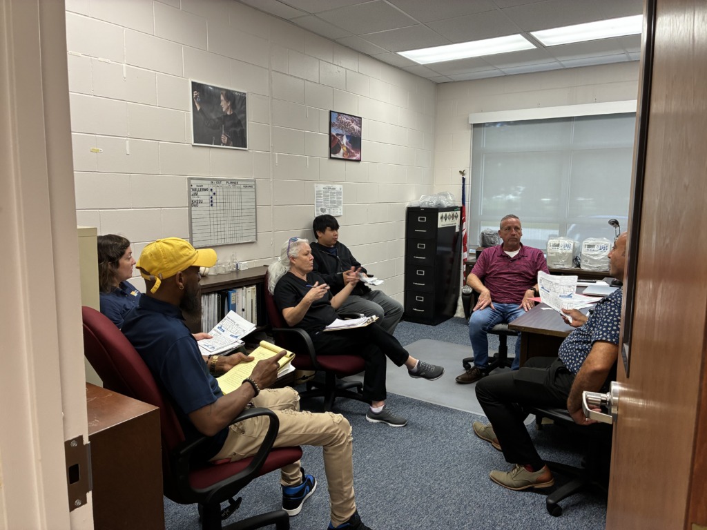 Seven people are seated in an office meeting room, actively discussing. One man in a yellow cap points at a notepad, while others listen and gesture. The room has cinder block walls, office furniture, and an 'IN/OUT PLANNER' whiteboard.