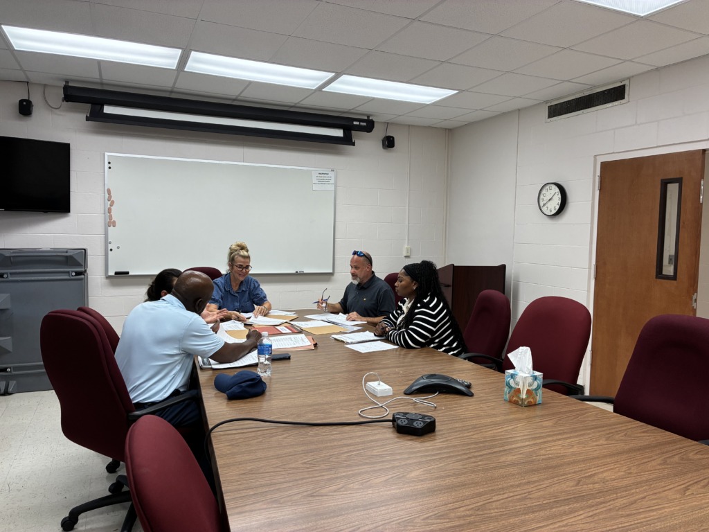 Five people, three men and two women, sit around a large wooden conference table, engaged in a meeting with papers and folders spread out. A whiteboard, TV, and clock are visible in the background.