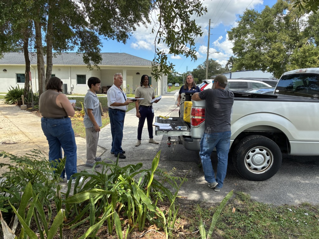 Six people gathered outdoors around a pickup truck with its tailgate down, discussing papers on a sunny day.