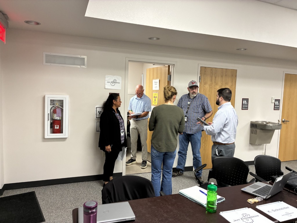 Five people, including men and women, standing in an office hallway or meeting area, engaged in conversation. Signs for 'Jaff Engineering,' 'Storage Room,' and 'Restroom' are visible on the walls, along with a fire extinguisher and a water fountain. A table with laptops and notebooks is in the foreground.
