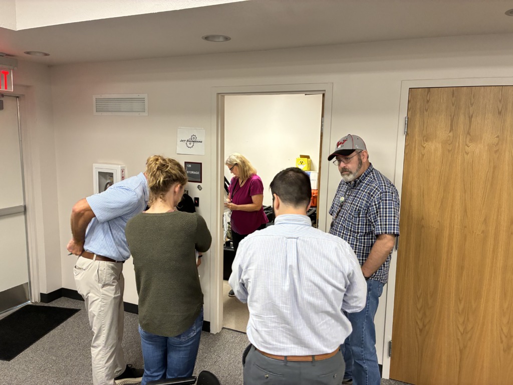 Five people, including a woman in a magenta shirt and a man in a plaid shirt and baseball cap, gather around a doorway labeled 'JAFF ENGINEERING 139 STORAGE' in an office setting.