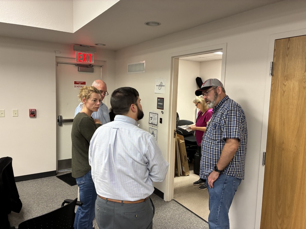 Five people, three men and two women, are gathered in an office hallway, engaged in conversation. An illuminated red 'EXIT' sign is visible above a door in the background.