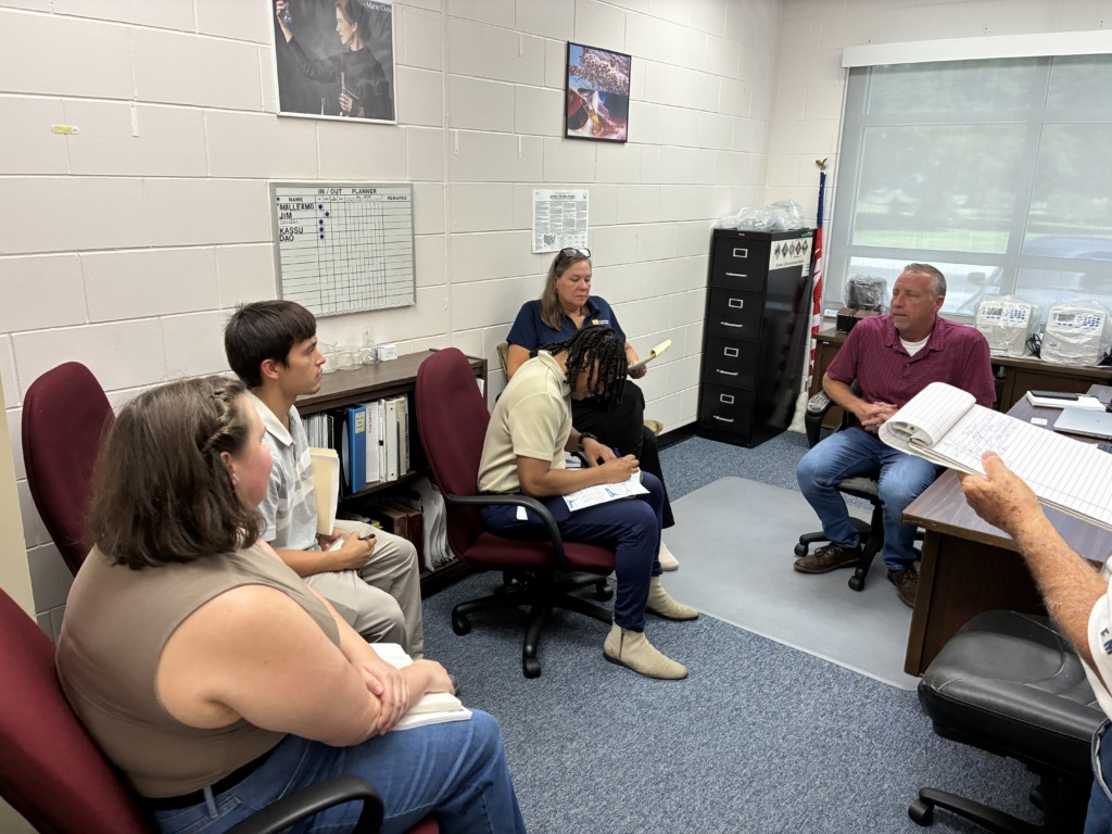 Six people, including adults and younger individuals, are seated in an office, engaged in a meeting or discussion. They are surrounded by office furniture, wall decor, and a window.