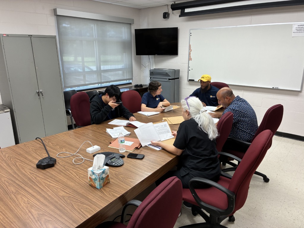 Five people of diverse backgrounds are seated around a conference table, reviewing documents and engaging in discussion. One person is on a phone call. The room features a whiteboard, TV, and window.