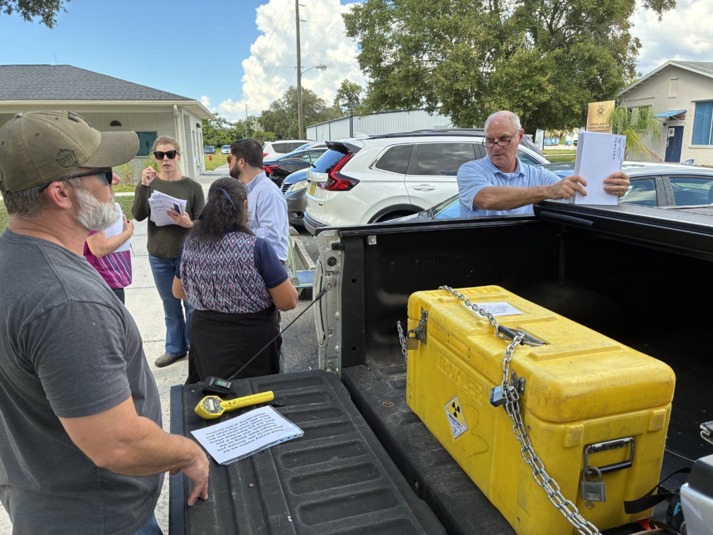A group of people, including men and women, gathered around the open tailgate of a pickup truck. In the truck bed, a bright yellow container with a 'RADIOACTIVE I' label is secured with chains, alongside a yellow handheld survey meter and instructions. People are holding papers and discussing, suggesting a training or inspection event.