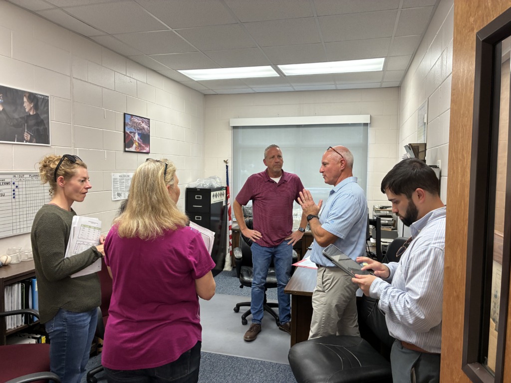 Five people, three men and two women, discussing in a brightly lit office, with one man gesturing as he speaks and others listening or looking at devices.