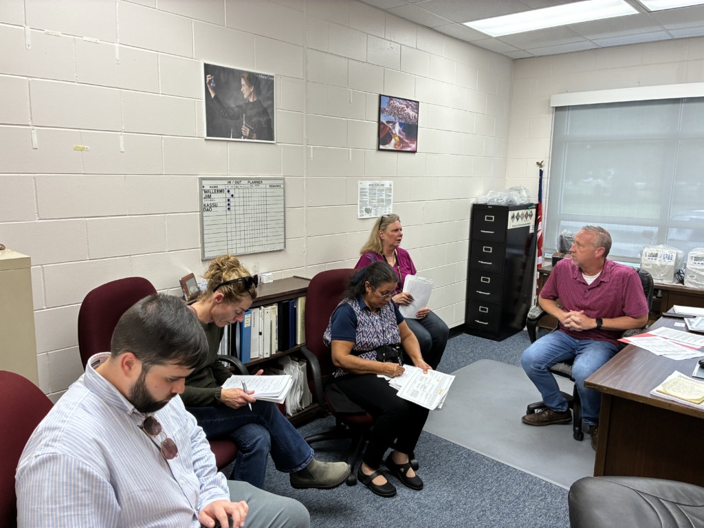 Five people are seated in an office, some taking notes, during a meeting. A whiteboard and a Marie Curie poster are visible on the wall.