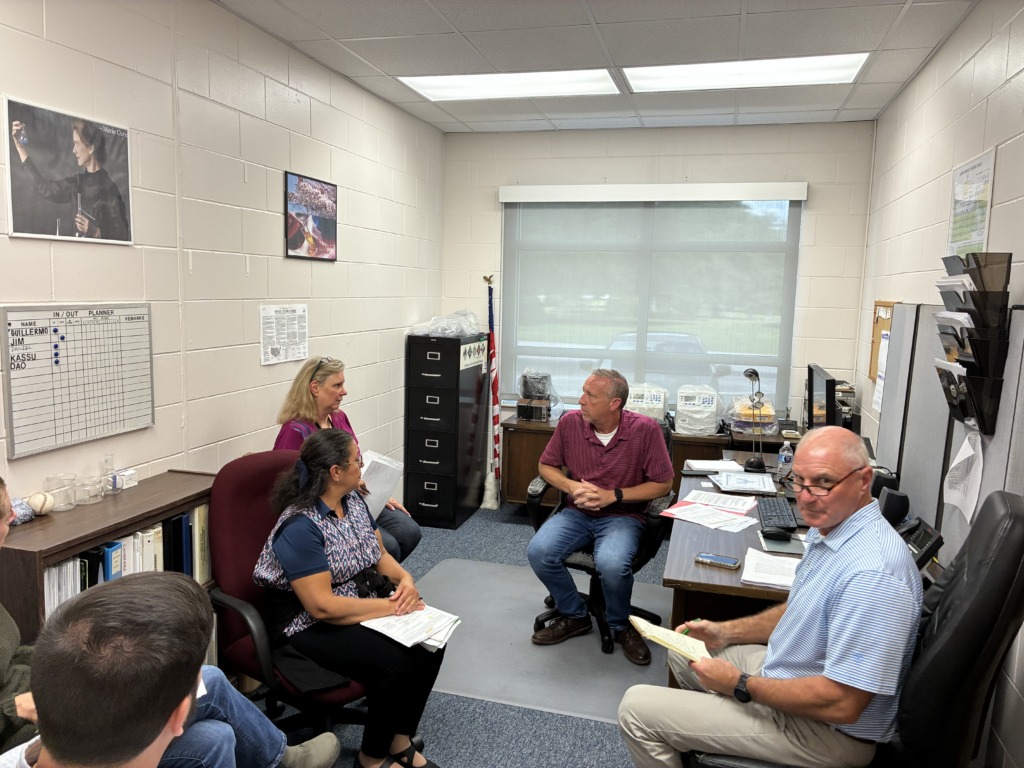 Five people are gathered in a small office for a meeting. A man in a red shirt sits at a desk, while others, including two women and another man, listen and take notes. An American flag and a Marie Curie poster are visible in the background.