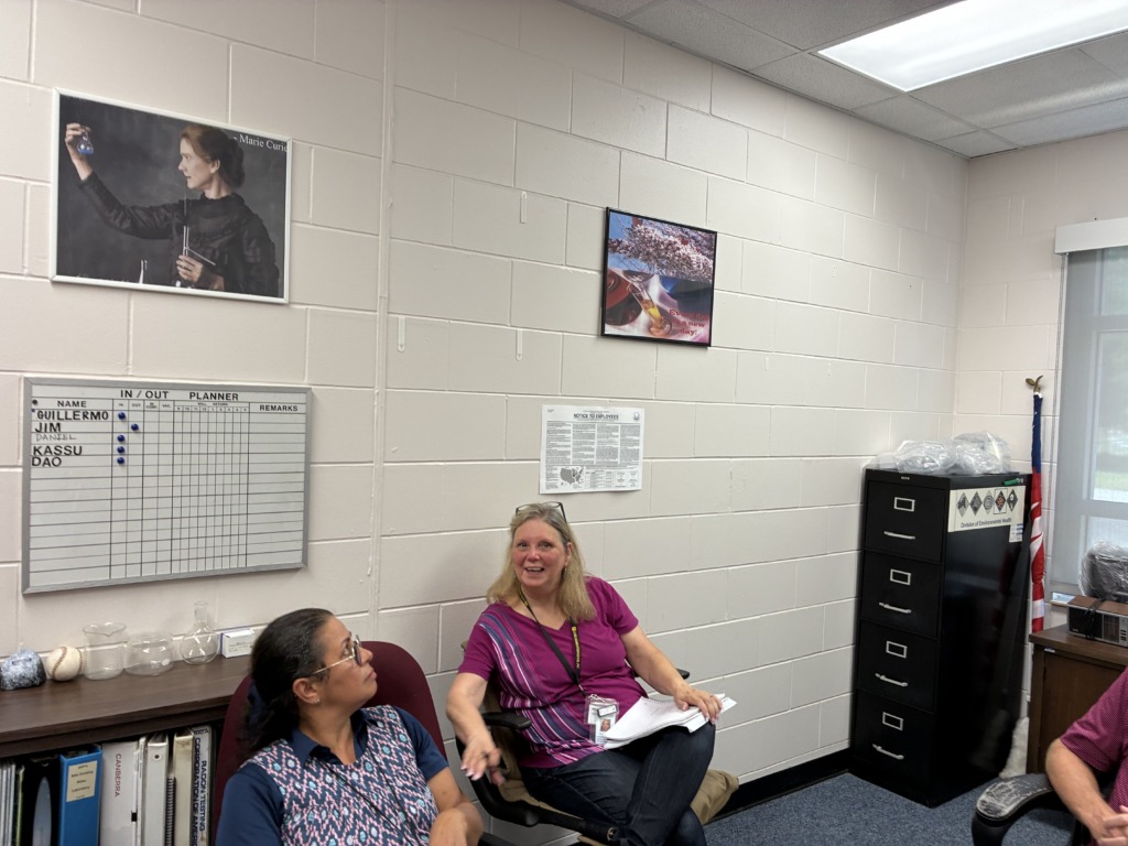Two women talk in an office with white cinder block walls. A Marie Curie poster, an 'in/out' board, and scientific glassware are visible on shelves and walls. A filing cabinet and American flag are in the background.