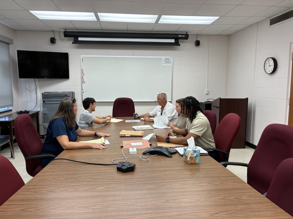 Five people are seated around a large wooden conference table in a meeting room, reviewing documents and discussing.