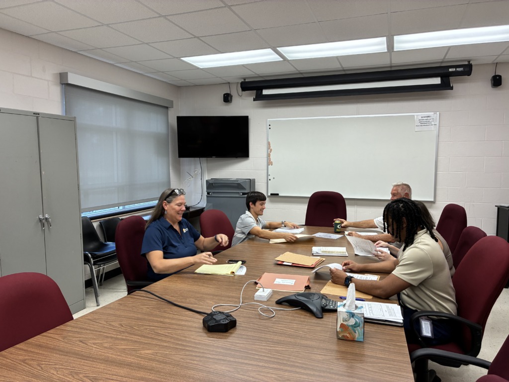 Four people, two men and two women, are seated around a large wooden conference table, reviewing documents and engaging in discussion. The room has white walls, a TV, a whiteboard, and a window with a blind.