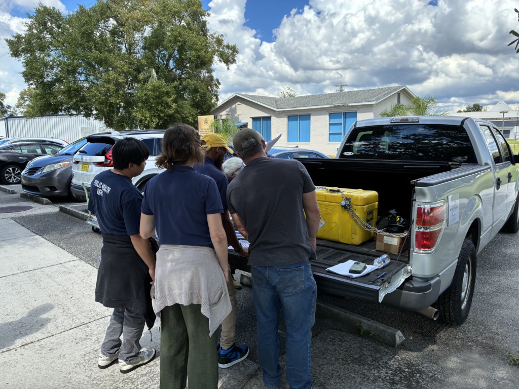 Five people, including one wearing a 'PUBLIC HEALTH' shirt, gather around the open tailgate of a silver pickup truck, reviewing papers next to a chained yellow container with a radiation symbol. A building and trees are in the background.