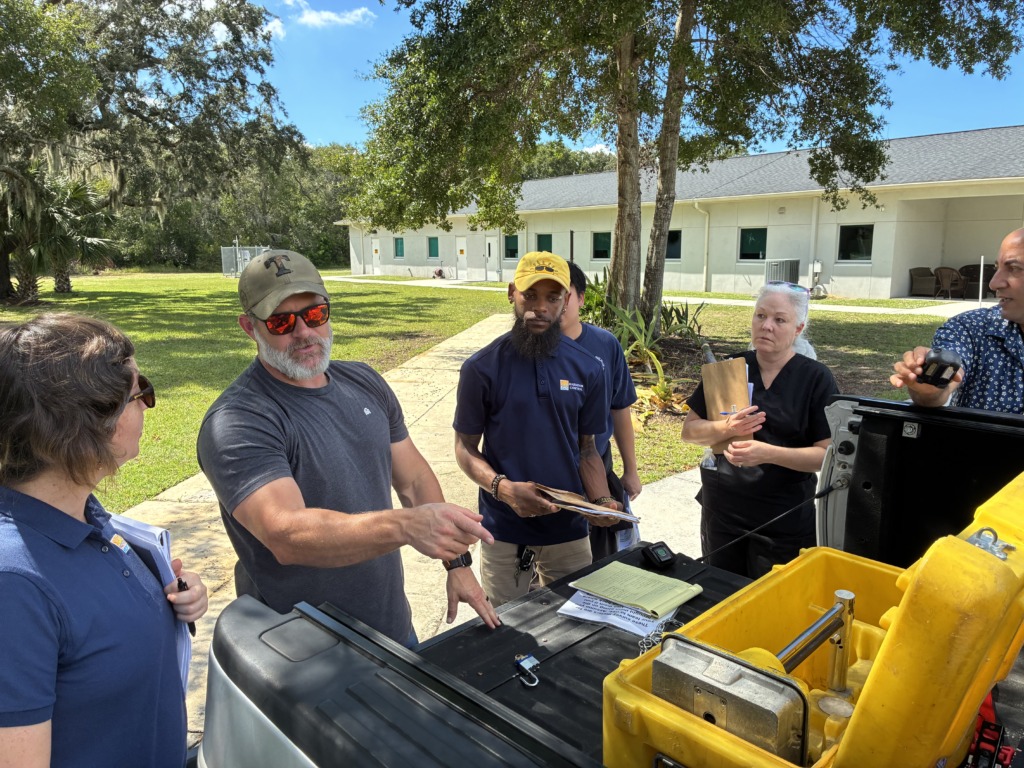 A group of five professionals, including a man in a 'Radiation Control' shirt, gather around the open tailgate of a pickup truck, discussing equipment in a yellow case on a sunny day outdoors.