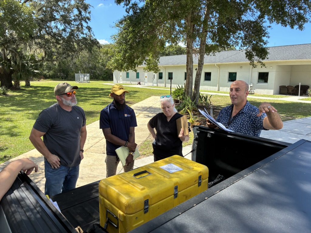Four individuals gathered around the open bed of a truck containing a yellow case labeled 'radioactive material,' with one person speaking and gesturing, in a sunny outdoor environment.