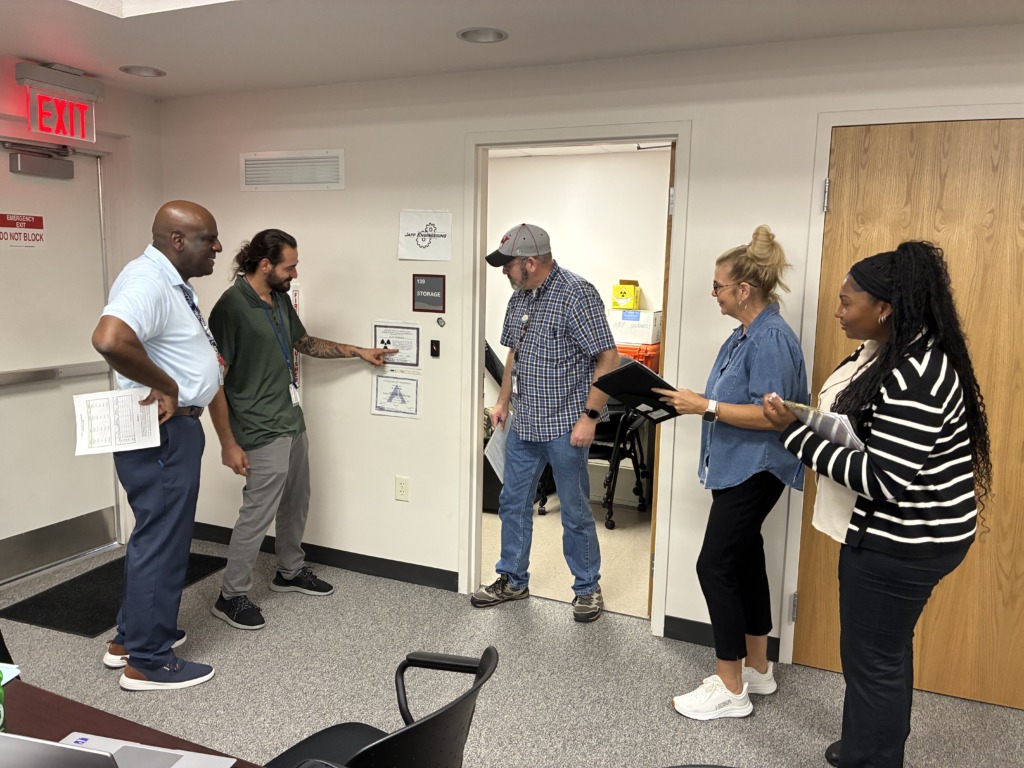 Five professionals, including men and women of diverse backgrounds, are gathered in a hallway, with one man pointing to a sign on the wall, possibly during an inspection or audit of a storage area.