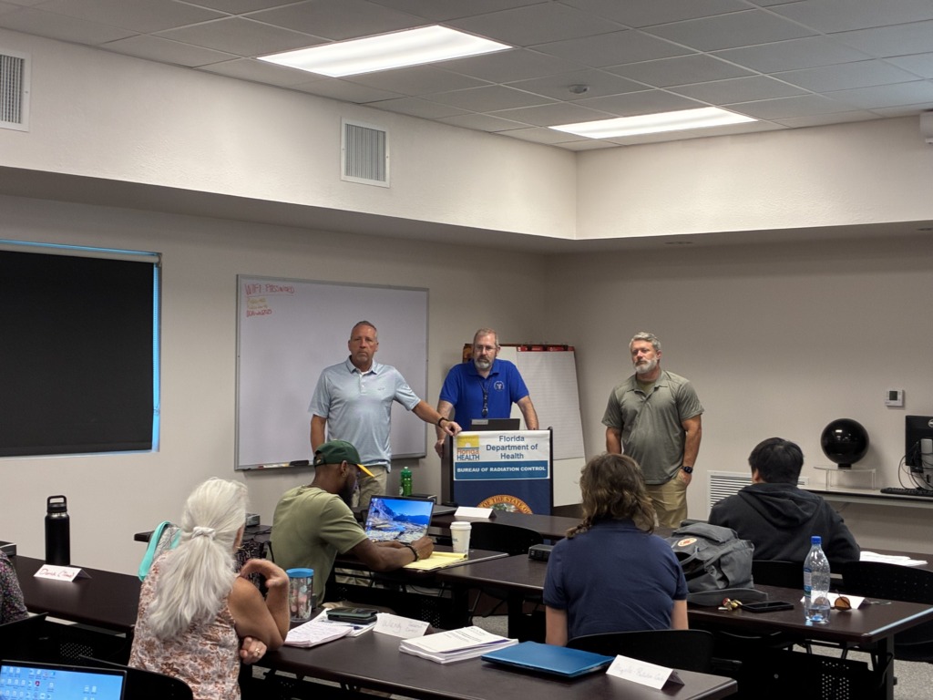 Three men from the Florida Department of Health, Bureau of Radiation Control, present to a group of attendees seated at tables in a brightly lit classroom.