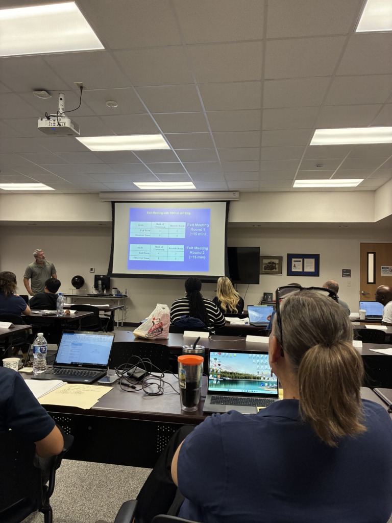 A classroom or conference room scene with attendees facing a projector screen displaying an "Exit Meeting" schedule. A person with brown hair in a ponytail is visible from behind in the foreground, with a laptop open on the table.