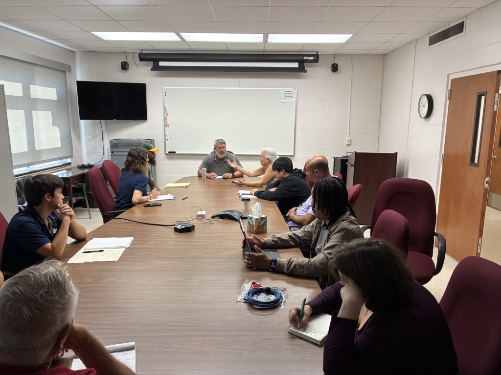 A diverse group of ten people seated around a large conference table in a meeting room, engaged in discussion. Papers, notebooks, and devices are on the table. A whiteboard, TV, and clock are visible on the walls.