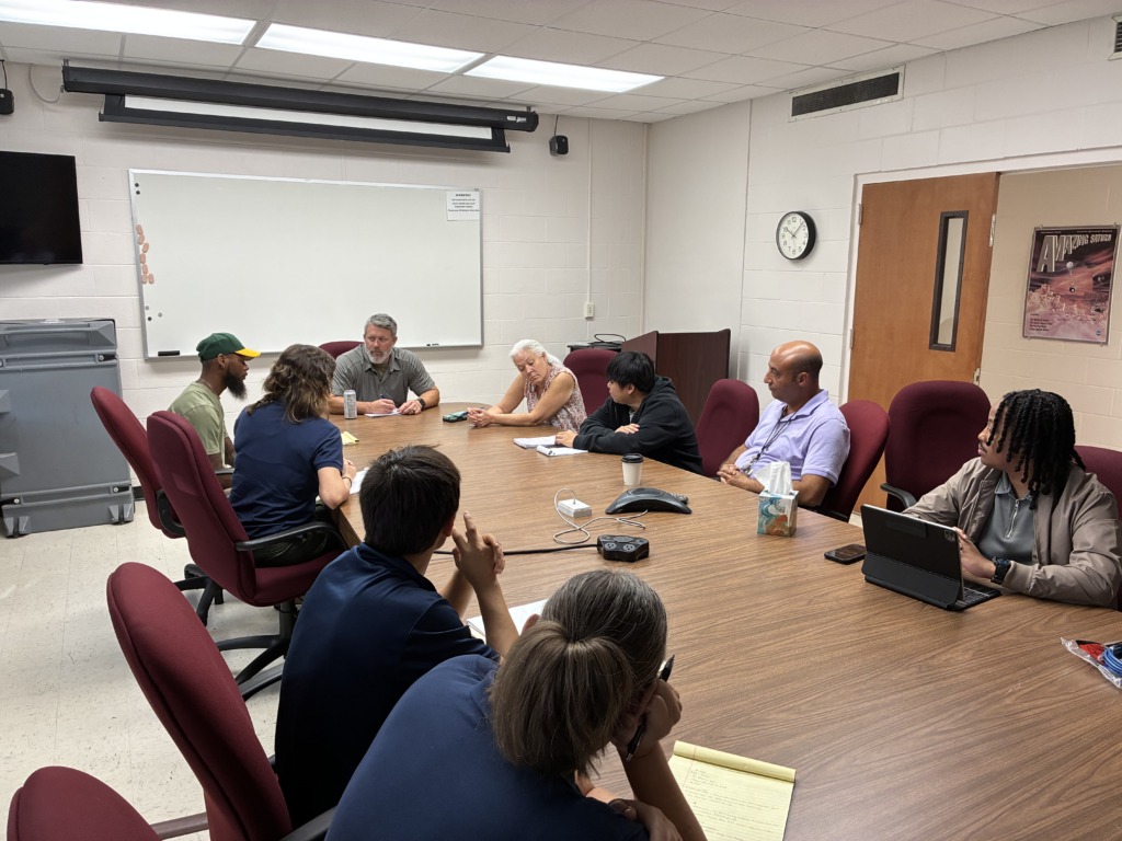 A group of diverse individuals seated around a large wooden conference table, engaged in a meeting or discussion.