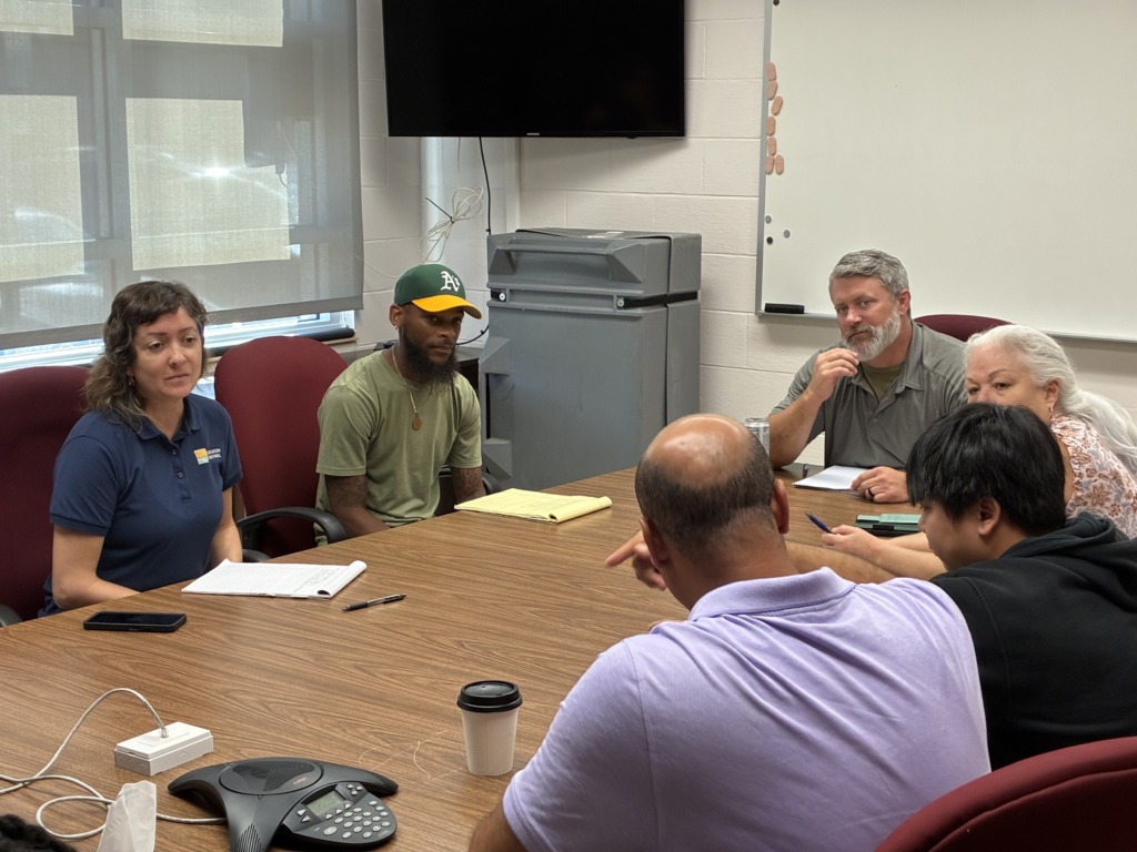 Seven people of diverse ages and backgrounds are seated around a conference table, engaged in a meeting. Some are looking at each other, others are taking notes. A conference phone and coffee cup are on the table.