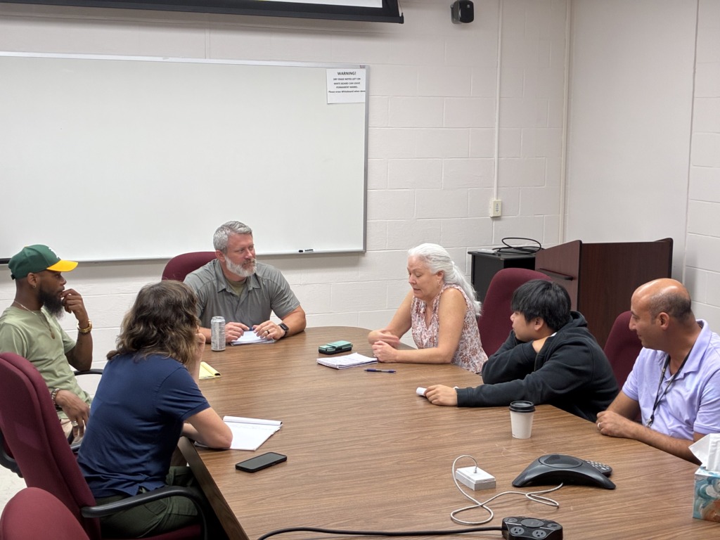Six people of diverse ages and backgrounds sit around a wooden conference table, engaged in a discussion. A whiteboard is in the background.