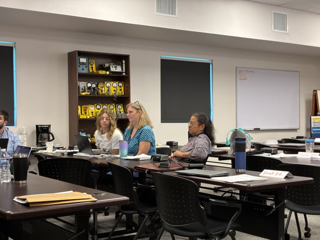 Three people, primarily women, seated at tables with laptops in a classroom or meeting room. A bookshelf filled with vintage electronic equipment is in the background.