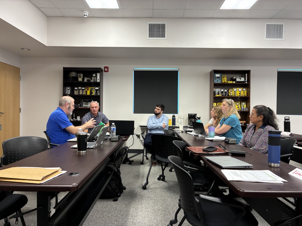 Six people, a mix of men and women, are seated around a long conference table with laptops, engaged in a discussion in a modern meeting room.