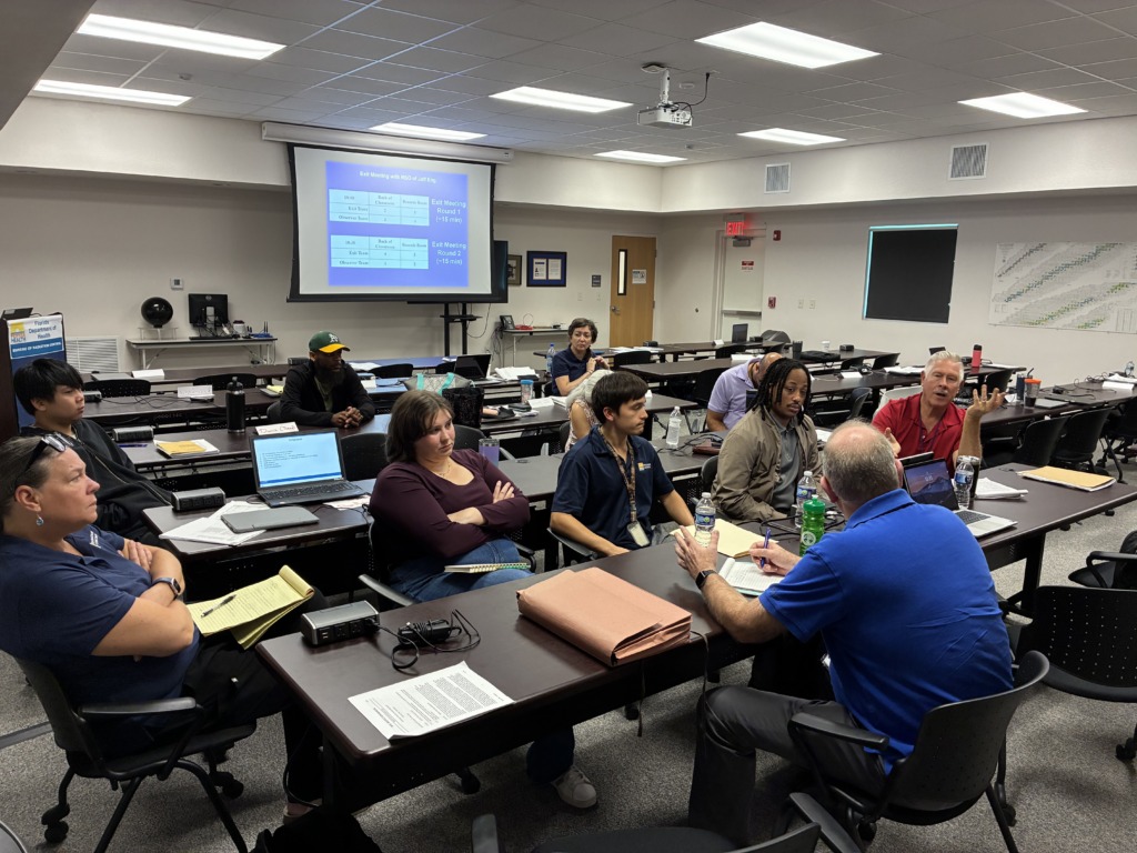 A group of diverse professionals, including men and women, are seated around tables in a classroom, engaged in a meeting. A projector screen displays a meeting schedule, and some individuals are taking notes or speaking.
