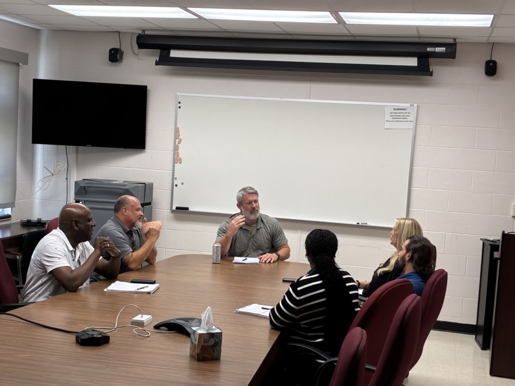 Six people, a diverse group of men and women, are seated around a large wooden conference table in a meeting room, actively engaged in discussion. A whiteboard and TV are visible in the background.
