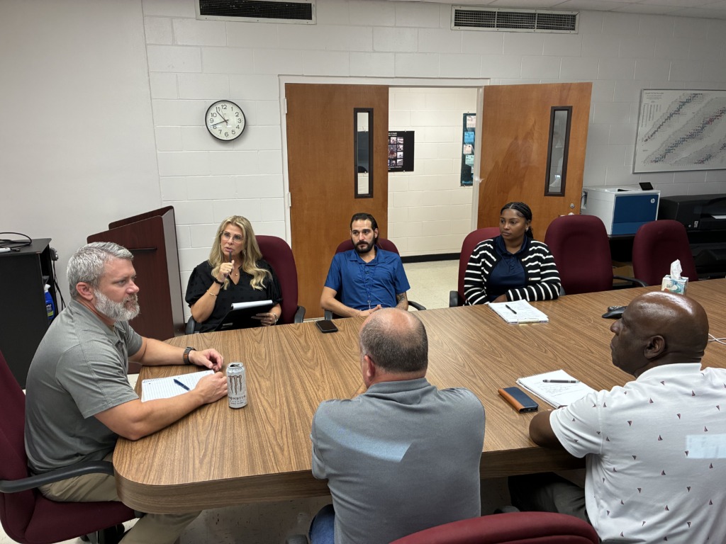 A diverse group of six people, including men and women, are seated around a large wooden conference table in a meeting room, engaged in discussion. Some are taking notes, and one woman appears to be speaking.