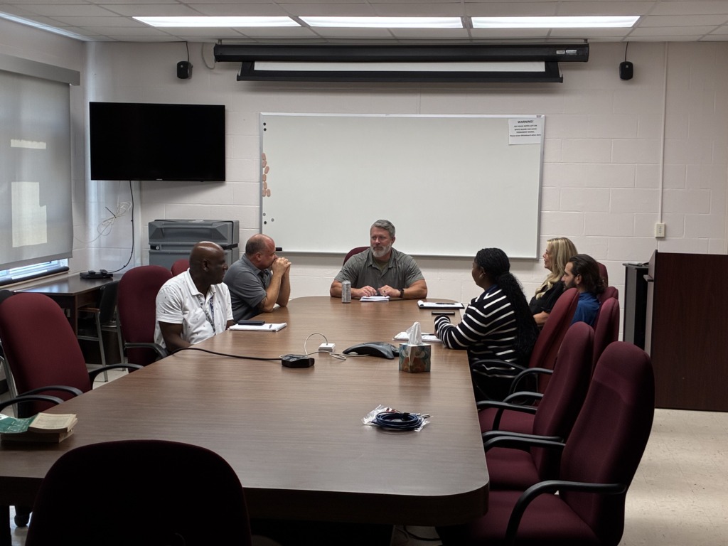Six people of diverse backgrounds sit around a large wooden conference table in a meeting room, engaged in discussion. A whiteboard and TV are visible in the background.