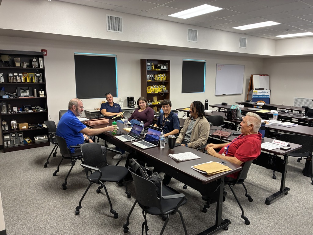Seven people, diverse in age and gender, sit around a long table in a classroom. Laptops, water bottles, and notebooks are on the table. Bookshelves filled with scientific or radiation detection equipment line the left wall, and a sign for the Florida Department of Health is visible on the right.
