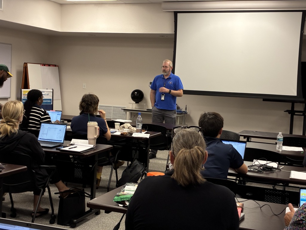 A man in a blue shirt stands in front of a blank projection screen, speaking to a group of people seated at tables with laptops. A whiteboard is visible on the left.