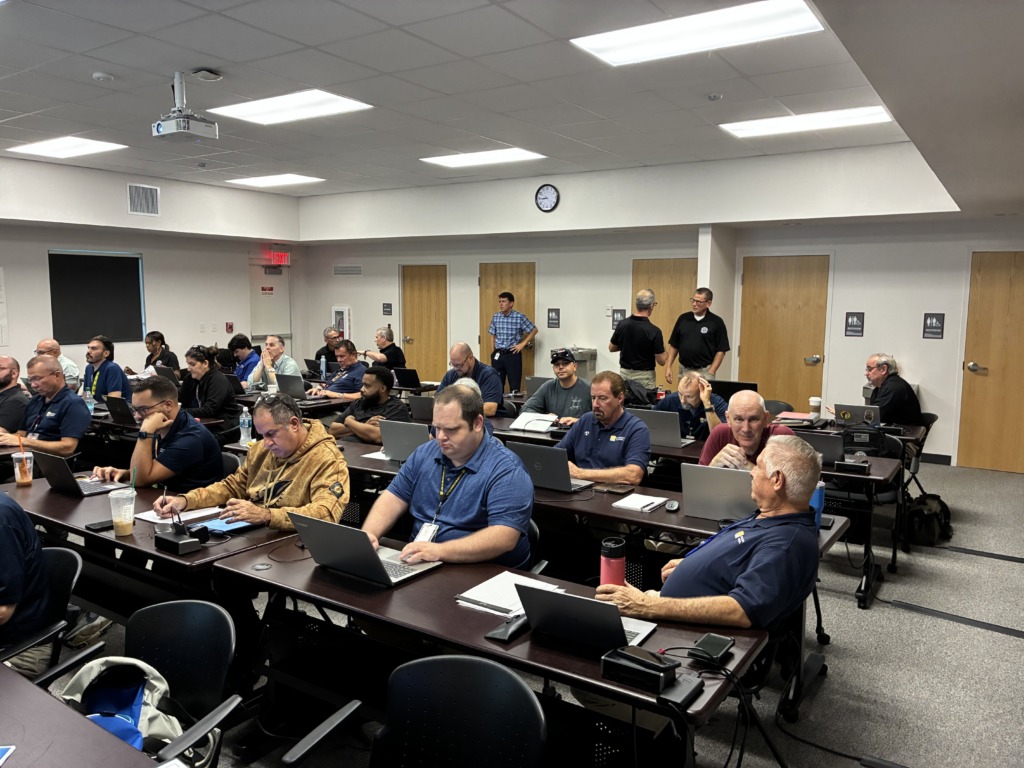 A classroom filled with about two dozen adults, mostly men, seated at individual desks, actively using laptops and taking notes during a training session. A projector is visible on the ceiling.