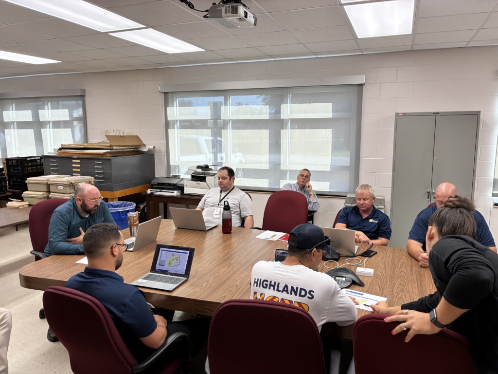 Eight people sitting around a large wooden conference table in a meeting room, looking at laptops and papers, with a map visible on one laptop screen.